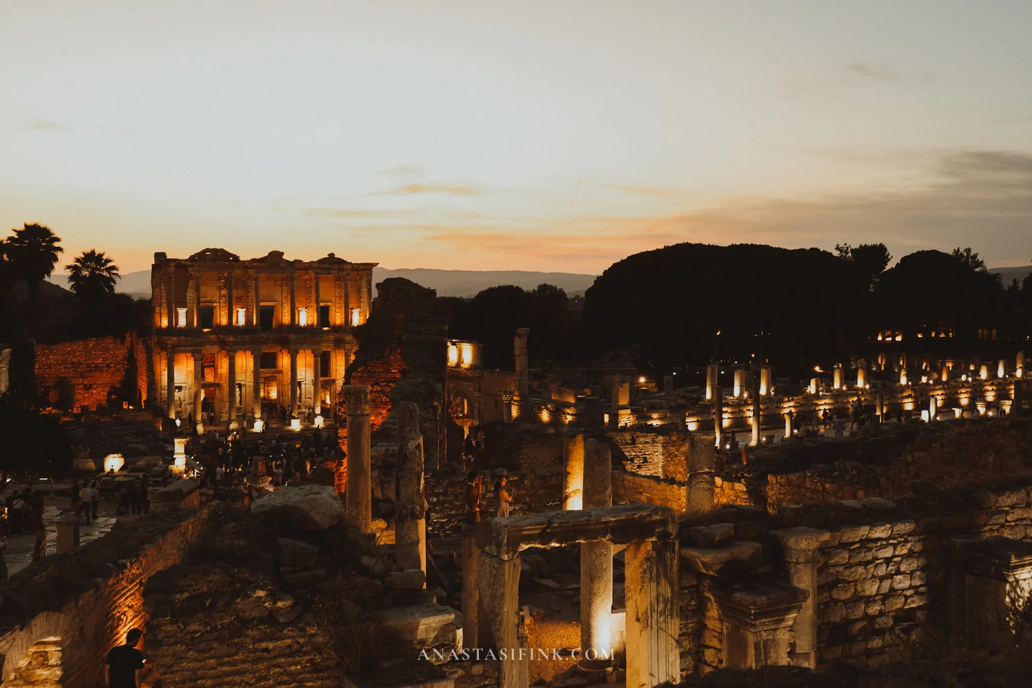 Wide night panorama of Ephesus — the Library of Celsus glowing amber, columns lit along the street