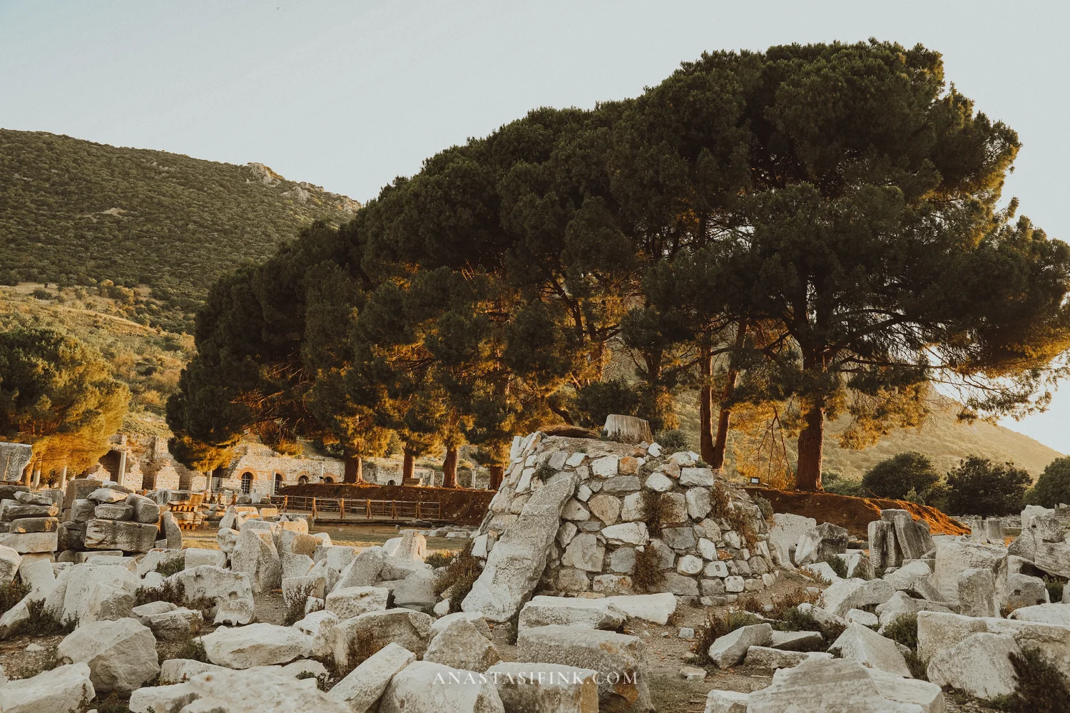 Ruins with tall pine trees against mountains, scattered marble blocks