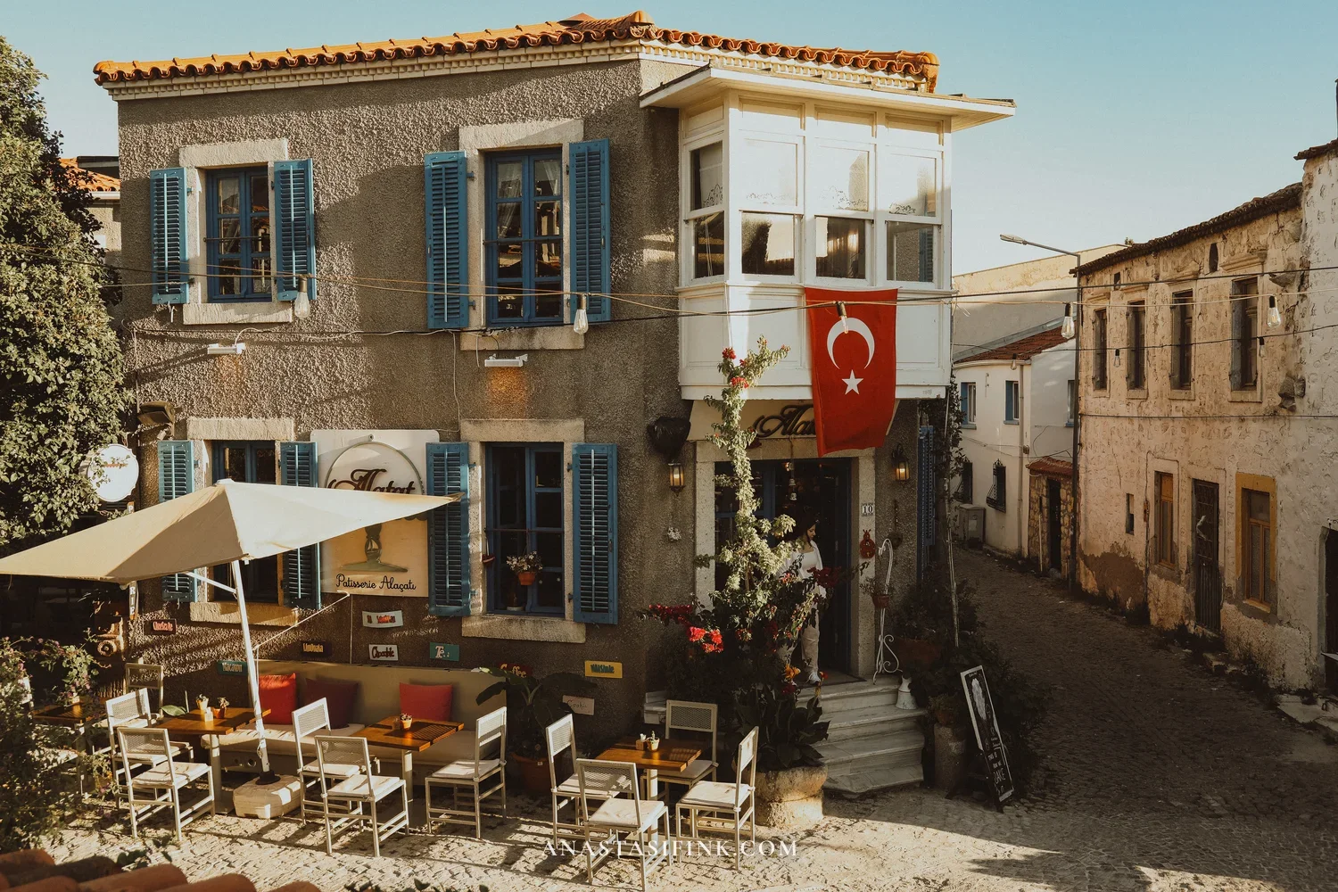 Café with blue shutters on an Alaçatı street — tables on the cobblestones, stone walls