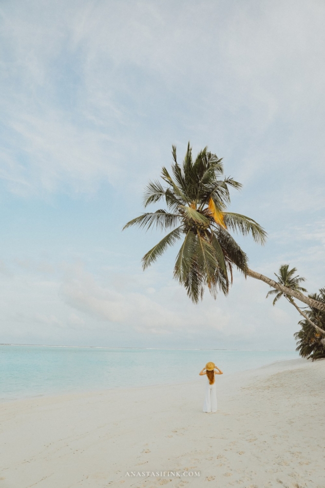 Woman in a white dress and straw hat under a leaning palm tree on a Maldives beach