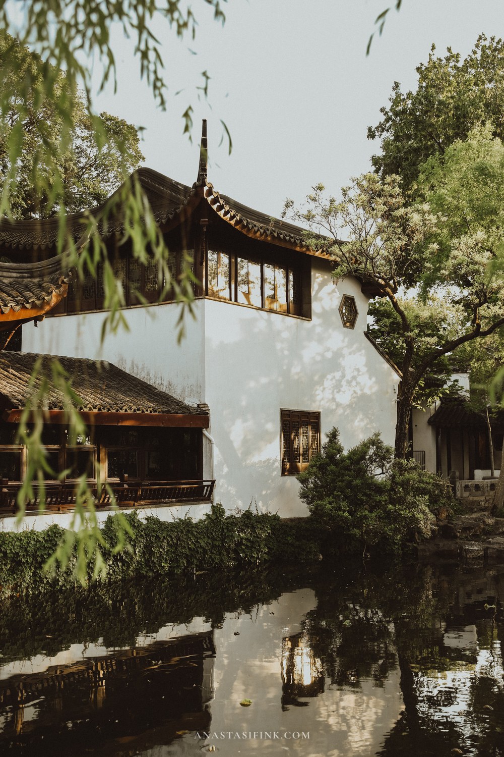 Traditional white garden hall with upturned tiled eaves seen through willow branches and mirrored in a pond