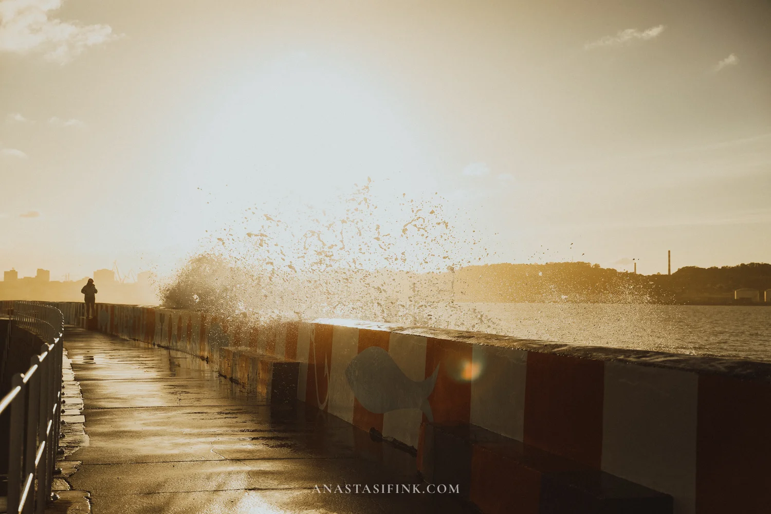 Wave crashing over the red and white breakwater wall in Gijón at sunset