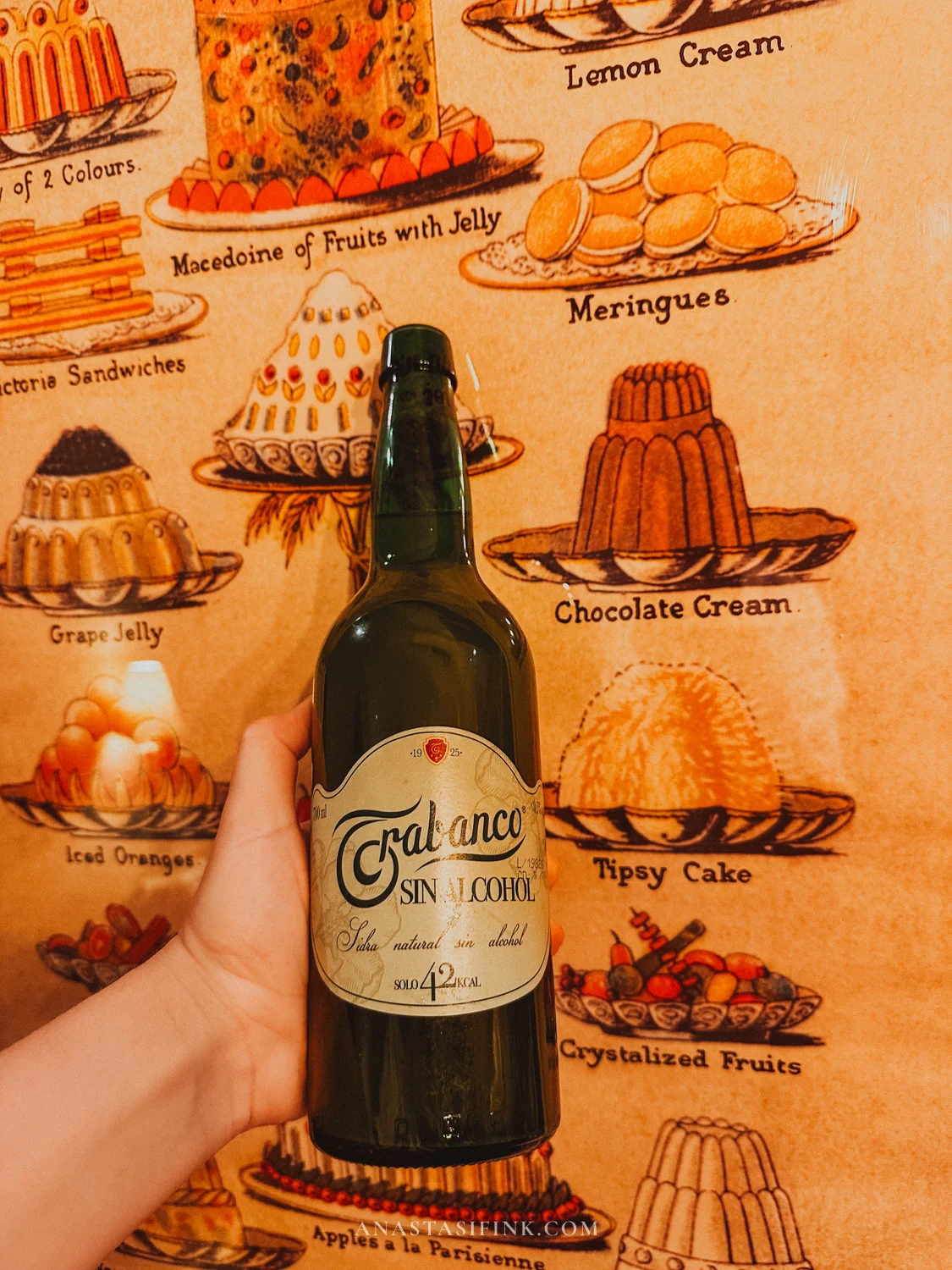 Bottle of Trabanco non-alcoholic natural cider held in front of a vintage dessert poster